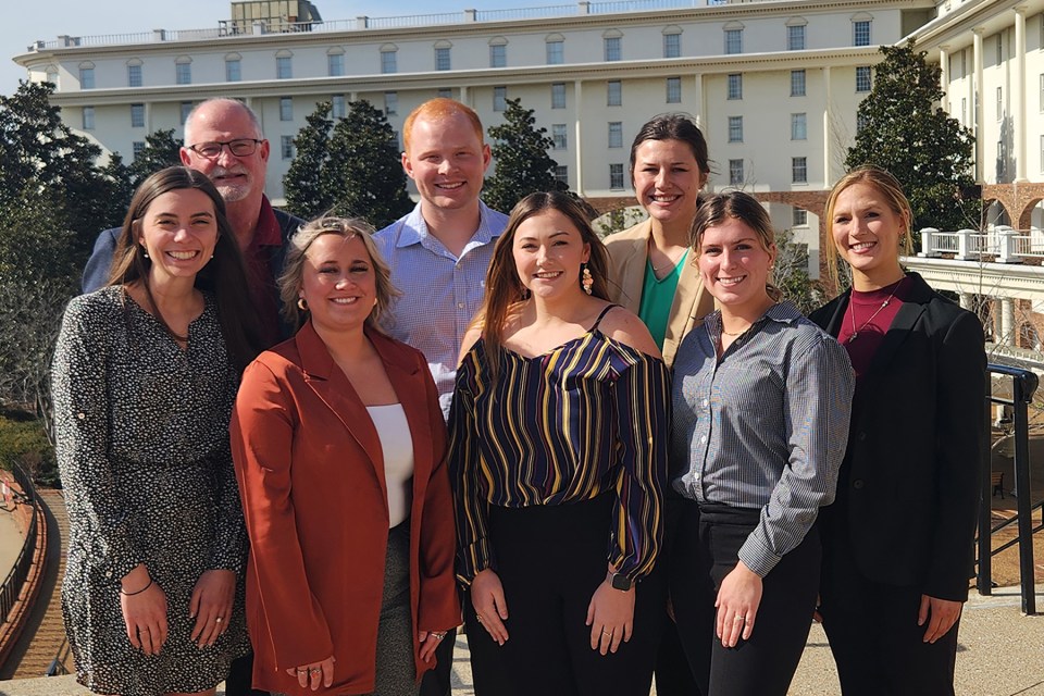 8 Veterinary medical student scholarship winners standing together with trees and a large cream colored building behind them.
